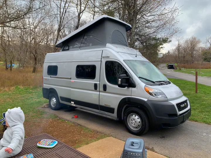 Silver Winnebago Solis pop-top camper van, an ideal RV rental, parked at a peaceful campground with a child enjoying outdoor play.. Winnebago Winnebago 2021