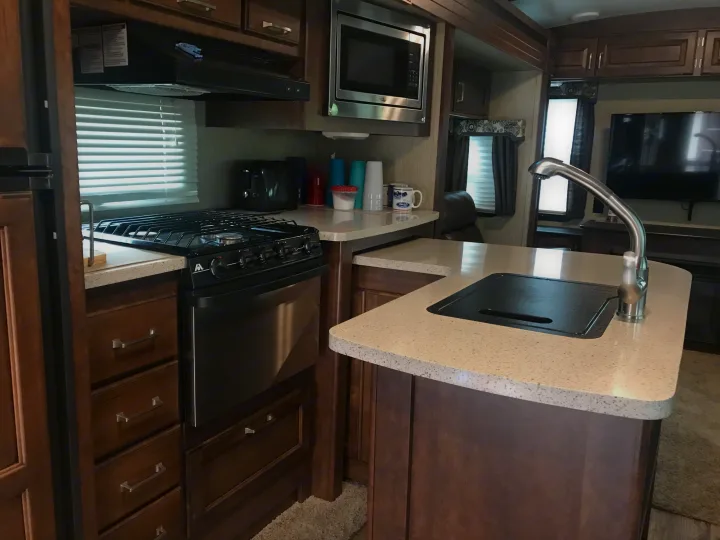 Modern RV rental kitchen showcasing a gas range, microwave, sink, light counters, and dark wood cabinets.. Keystone Outback 2018