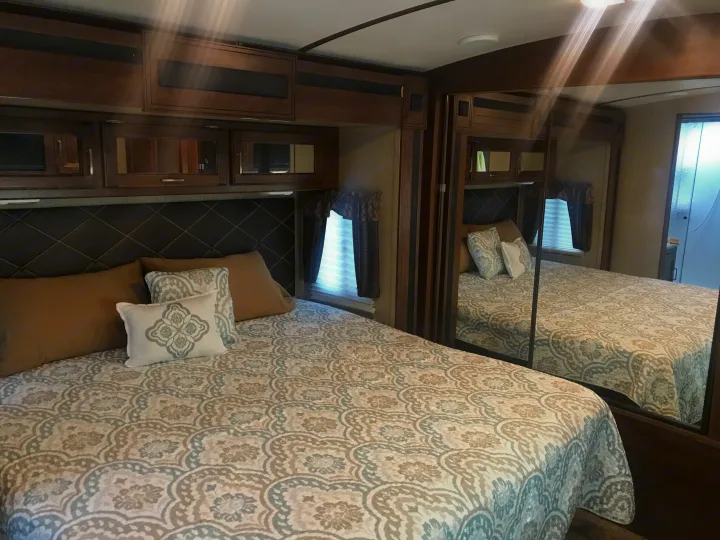 Cozy RV rental bedroom featuring a bed with elegant patterned bedding, dark wood cabinets, and a large mirrored closet.. Keystone Outback 2018