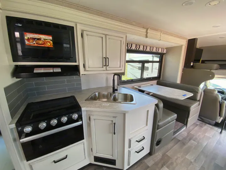 Inside an RV rental: a bright kitchen with white cabinets, microwave, stove, sink, and grey dinette, offering travel comfort.. Jayco Redhawk 2025