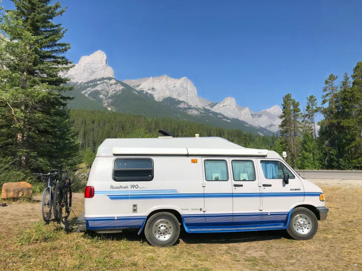 Roadtrip ready: an RV rental with bikes parked against a stunning mountain range and forest under a clear blue sky.. Roadtrek 190 Popular 1994