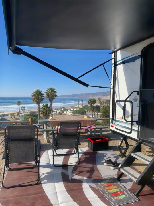Enjoying an RV rental view! Extended awning over outdoor chairs at a sunny, oceanfront campsite with palm trees.. Forest River Surveyor 2021