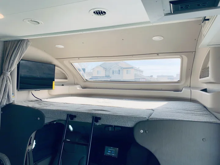 Explore the cozy over-cab bunk sleeping area of this RV rental, featuring a bed, large window, TV, and ladder access.. Entegra Coach Odyssey 31F 2021