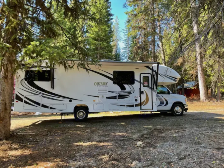 Explore the outdoors with an RV rental! A white Odyssey Integra motorhome is parked under trees at a sunny campground.. Entegra Coach Odyssey 31F 2021