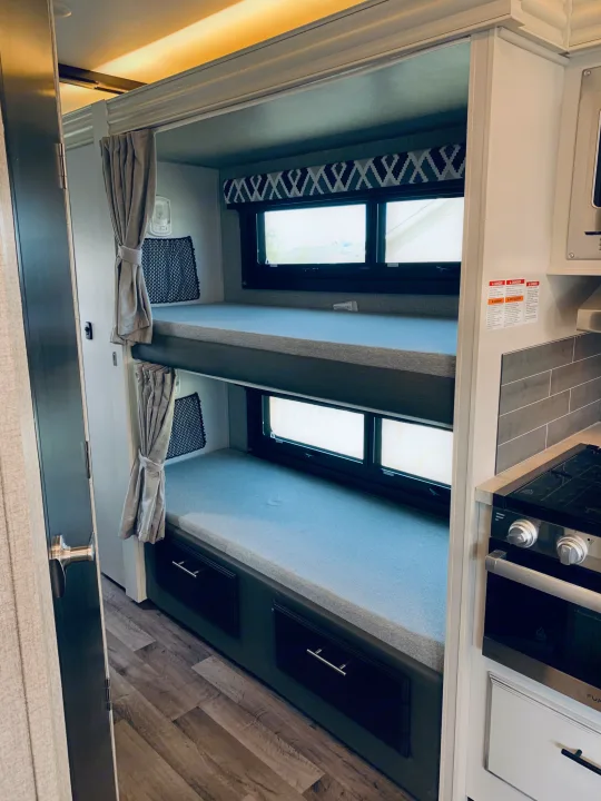 This RV rental bunk area offers cozy beds, windows, and storage. A compact kitchen with a stove is visible.. Entegra Coach Odyssey 31F 2021
