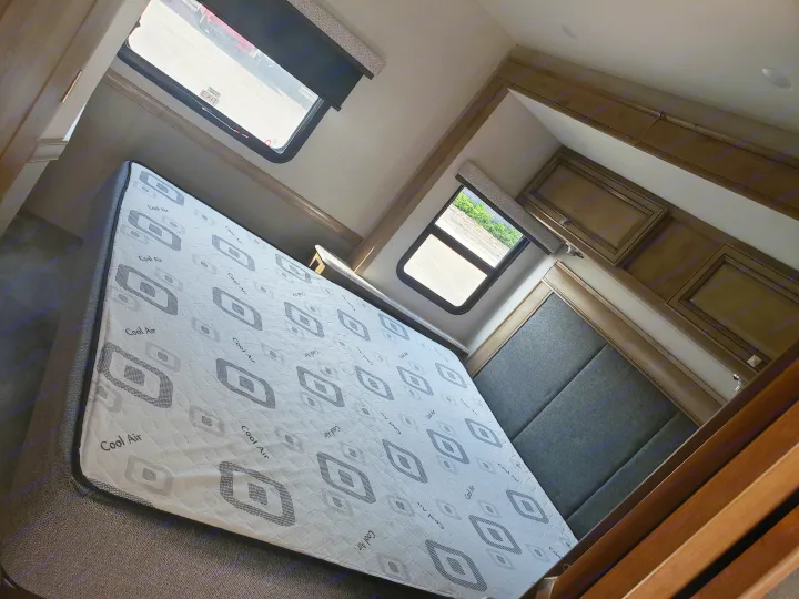 RV rental bedroom features a "Cool Air" mattress, dark headboard, and overhead cabinets. Windows show the exterior view.. Holiday Rambler Admiral 2021