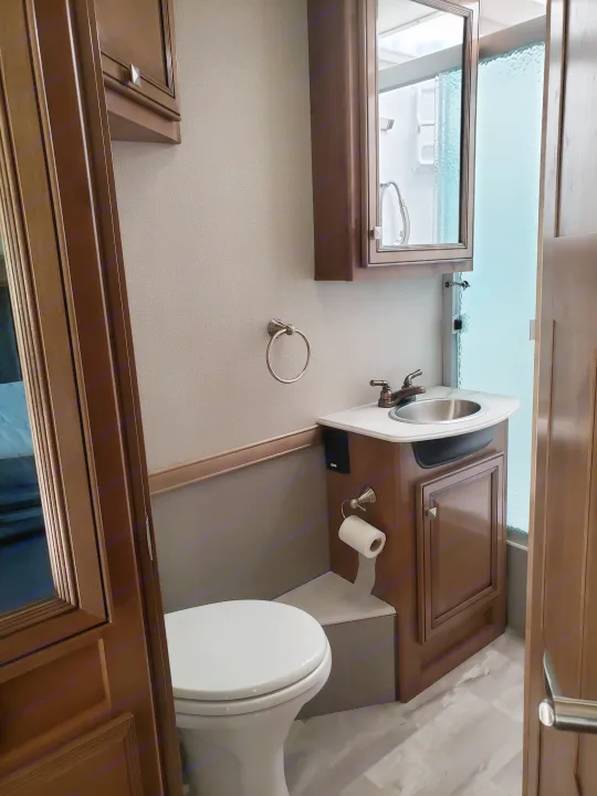 RV rental's compact bathroom: wooden vanity with sink, toilet, mirrored cabinet, and frosted glass shower stall.. Holiday Rambler Admiral 2021