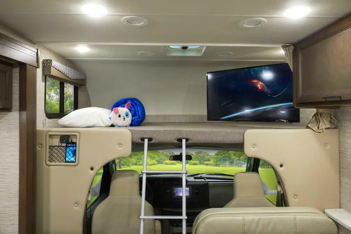 Bright RV rental interior showing a cab-over bunk with TV, pillow, stuffed toy, and ladder over the cabin.. Thor Freedom Elite 2021