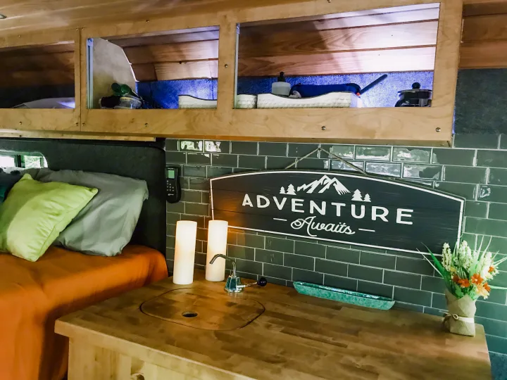 Cozy RV rental interior with kitchenette, bed, overhead storage, and an 'Adventure Awaits' sign. Ideal for trips.. Ram Promaster 1500 2021