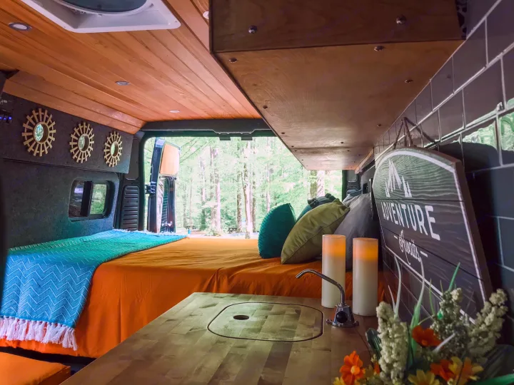 Cozy RV rental interior with an orange bed, blue blanket, wood ceiling, and modern sink counter in the foreground. Forest view.. Ram Promaster 1500 2021