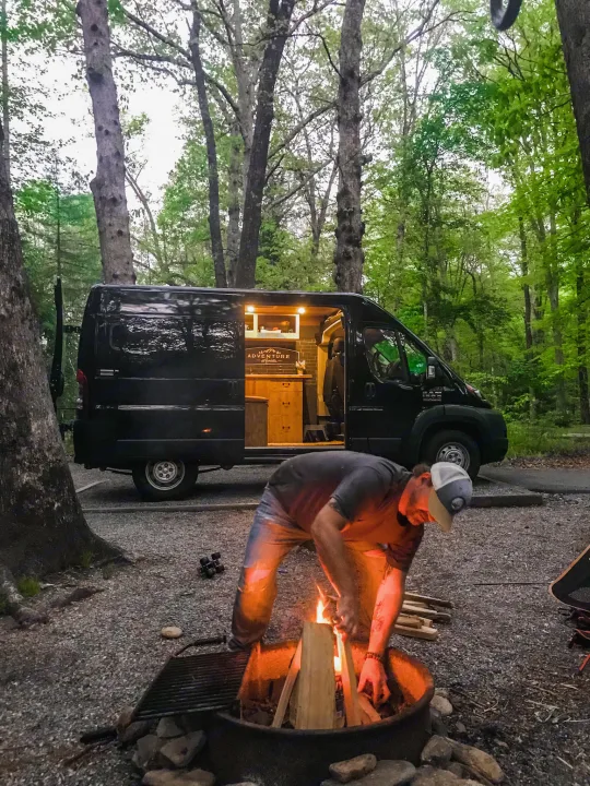 Evening camp vibes: A man kindles a campfire beside his cozy RV rental camper van at a peaceful wooded campsite.. Ram Promaster 1500 2021