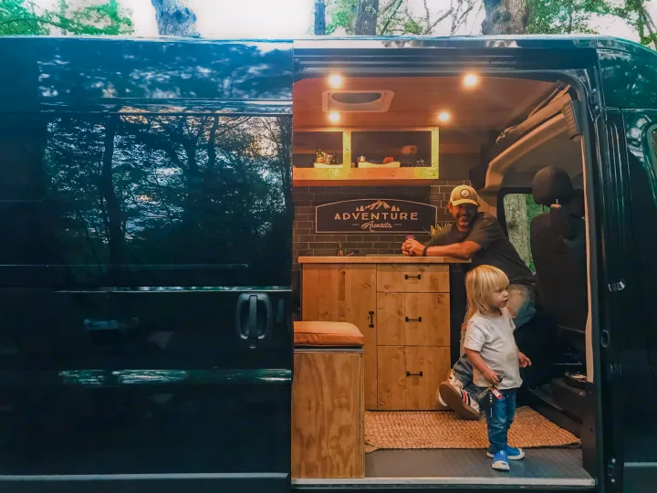 A man and child inside a custom RV rental camper van with a cozy wooden interior and "ADVENTURE Awaits" sign.. Ram Promaster 1500 2021