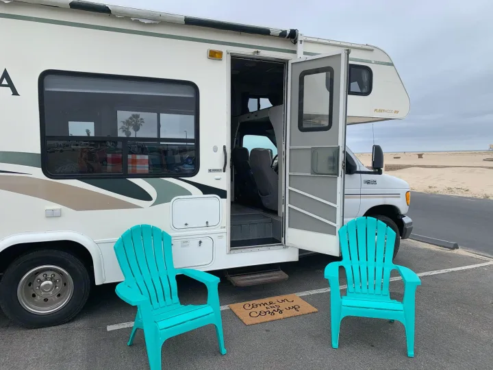 Beachside RV rental with open door, inviting teal chairs, and a 'Come in and Cozy up' mat. Perfect for outdoor living!. Fleetwood Tioga 2003