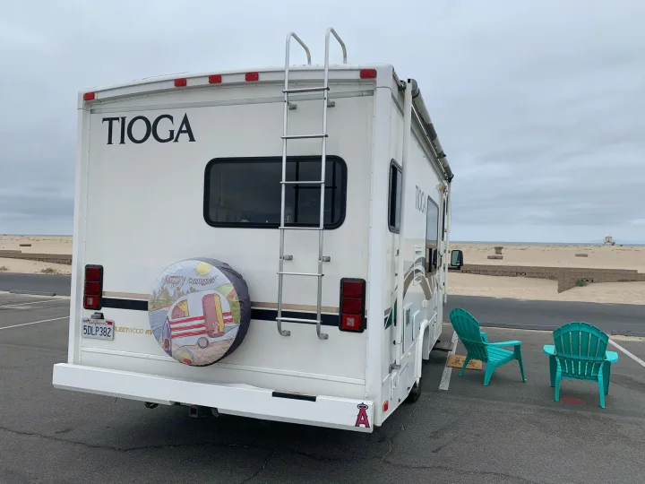 White RV rental parked near a sandy beach on a cloudy day, with two teal chairs beside it, ready for relaxation.. Fleetwood Tioga 2003