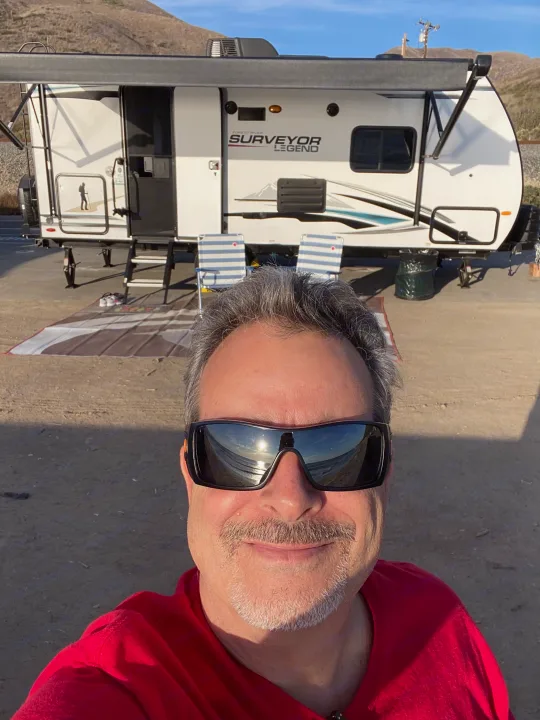Man smiles with sunglasses in front of his RV rental, a white Surveyor Legend travel trailer, under an awning.. Forest River Surveyor 2021