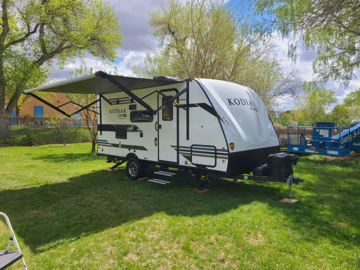 This RV rental, a white Kodiak Cub travel trailer, is set up on green grass with its awning extended under a partly cloudy sky.. Dutchmen Kodiak 2021