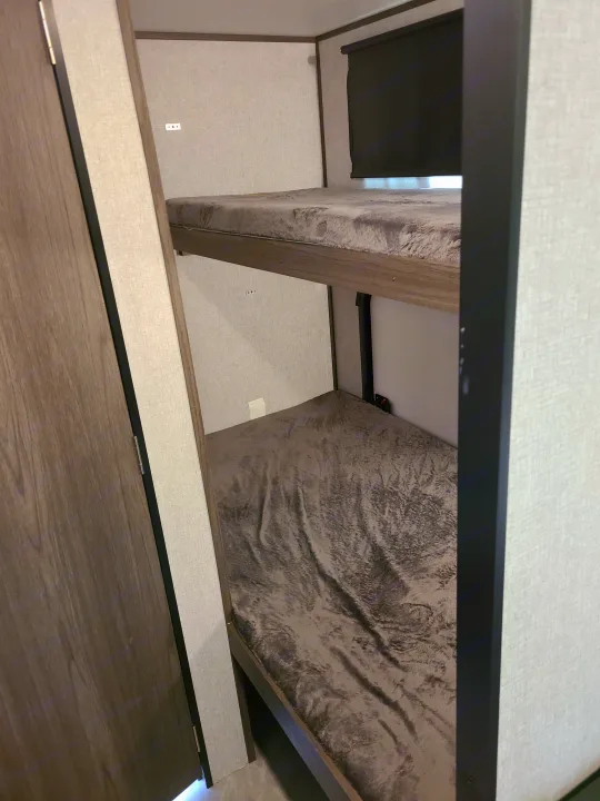 Cozy RV rental interior featuring two bunk beds with soft gray blankets and a privacy blind on the top bunk window.. Dutchmen Kodiak 2021