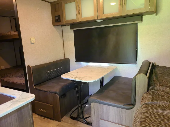 This RV rental features a cozy dinette with dark benches and a marble-patterned table, adjacent to bunk beds and a kitchen glimpse.. Dutchmen Kodiak 2021
