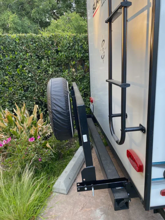 Rear of an RV rental, showing its white body, black ladder, and spare tire on a rack. Green hedges behind.. Forest River Surveyor 2021