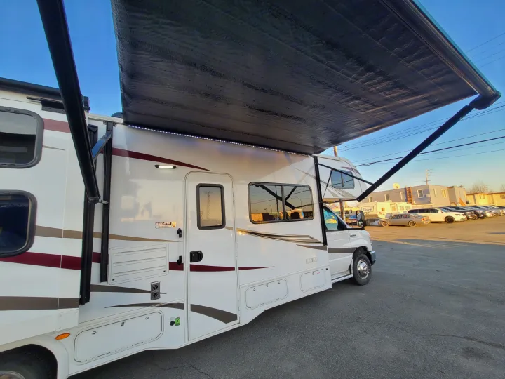 This modern white RV rental features a deployed awning with LED lights, parked outdoors on an asphalt lot.. Forest River Sunseeker 2018