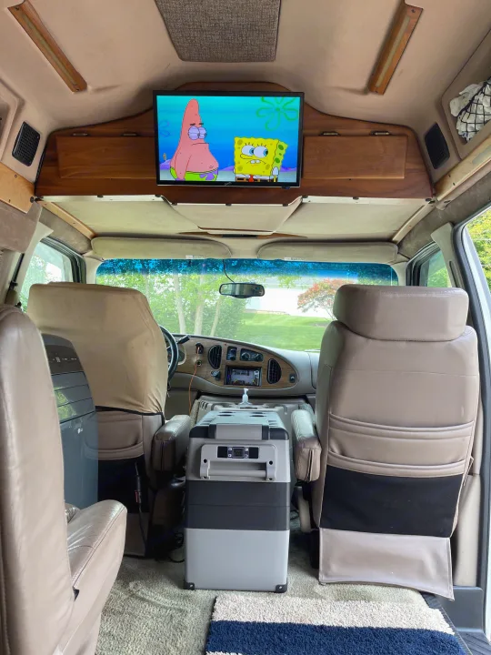 Experience cozy travel in this rv rental. Interior view shows overhead TV with cartoons, a portable cooler, and AC unit for comfort.. Coachmen Van Conversion 2001