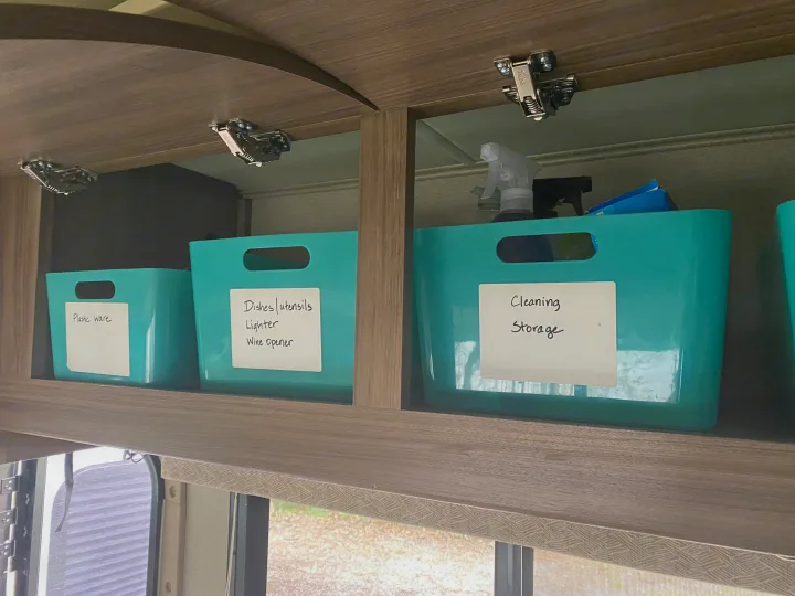 Organized RV rental interior. Turquoise storage bins labeled for plastic ware, dishes, and cleaning supplies in overhead cabinets.. Entegra Coach Qwest 2019
