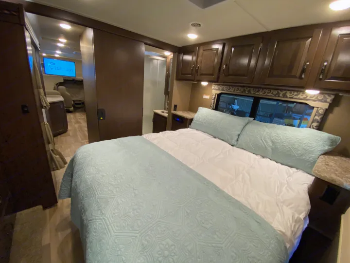 Comfortable RV rental bedroom with a large bed, dark wood cabinets, and a sliding door leading to the main living area with TV.. Thor Motor Coach Four Winds with full wall slide 2018