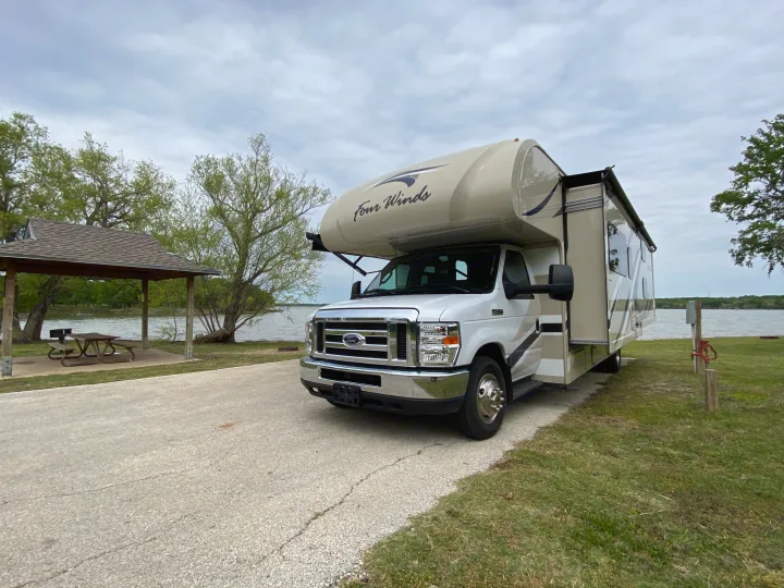 This RV rental, a Four Winds motorhome, is parked at a lakeside campsite with a picnic area.. Thor Motor Coach Four Winds with full wall slide 2018