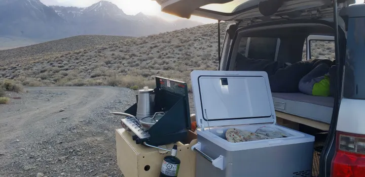 RV rental outdoor kitchen setup with camp stove, coffee, and fridge, beside a bed in a van, with mountains in background.. Honda Element 2003