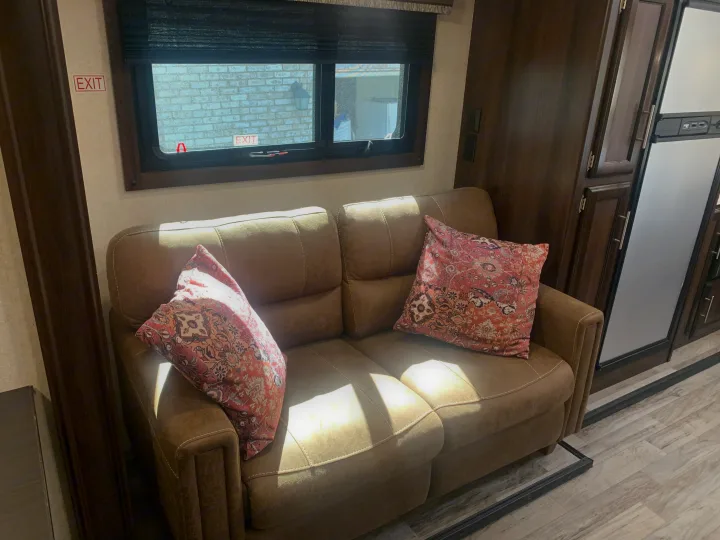Inside an RV rental: a comfortable brown sofa with two pillows, a window view, and dark wood cabinets/fridge.. Jayco White Hawk 2018