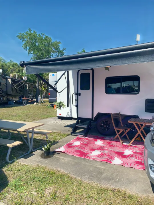 RV rental set up for comfort: White RV with extended black awning, red patterned rug, and picnic table at a sunny campsite.. Forest River no boundaries 2020