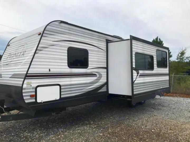 Side view of a gray & white Pioneer travel trailer with an extended slide-out, parked on gravel for RV rental.. Heartland Pioneer 2016