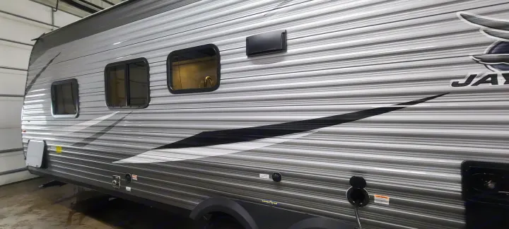 Side view of a corrugated gray and white Jayco RV rental, featuring windows, black accents, and utility hookups for travel.. Jayco Jay Flight 2021