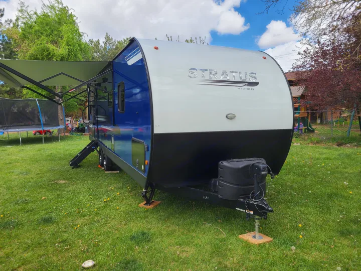 A blue and white Stratus RV rental, a travel trailer by Venture RV, with its awning extended, parked on a green lawn.. Venture Rv 261 VBH 2021