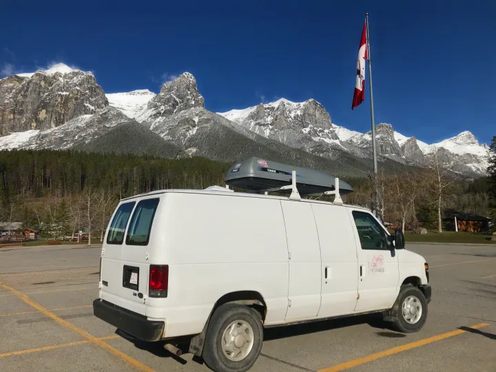 White RV rental van with a roof cargo box, parked in front of majestic snow-capped mountains and a Canadian flag.. Ford Econoline 2009