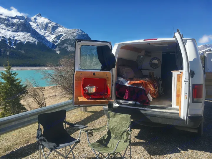Scenic rv rental adventure! Campervan with open doors reveals cooking setup & bed, overlooking turquoise lake & snowy mountains. Van life awaits!. Ford Econoline 2009