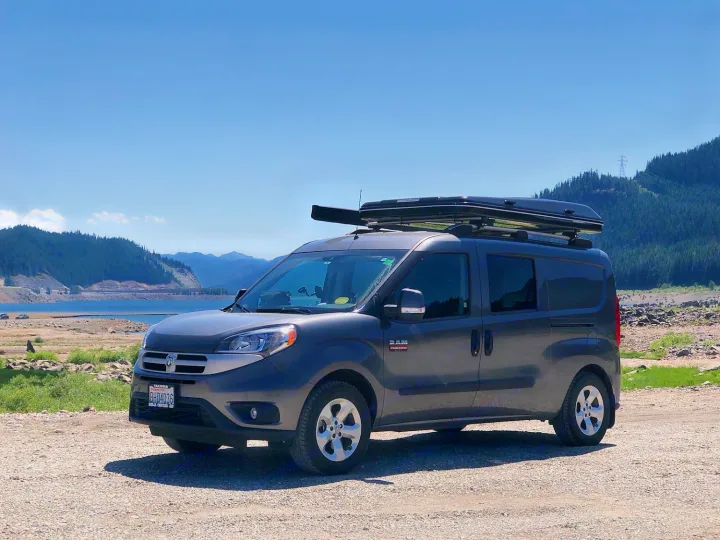Compact Ram ProMaster City van, set up as an RV rental with a rooftop tent, parked by a scenic lake for outdoor adventures.. Ram Promaster City 2021