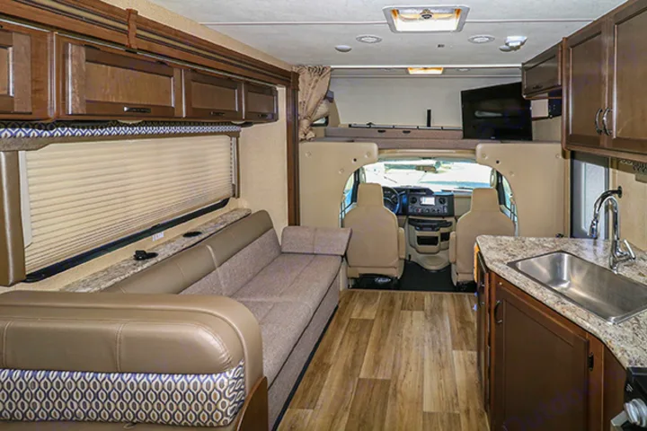 Inside a modern RV rental, featuring a living area with sofa, kitchen, driver's cabin, and overhead bunk bed with TV.. Thor Motor Coach Freedom Elite 2019