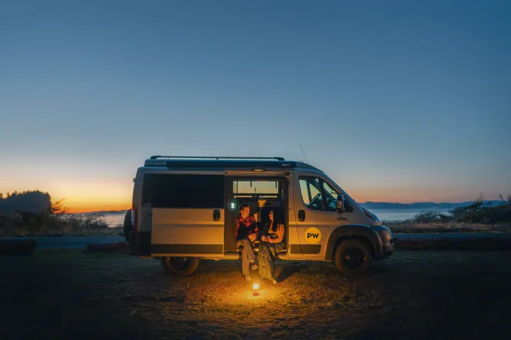 A cozy rv rental scene: A couple sits in their camper van, lit by a warm lantern, enjoying a beautiful sunset by the water.. Ram Pleasure Way 2019
