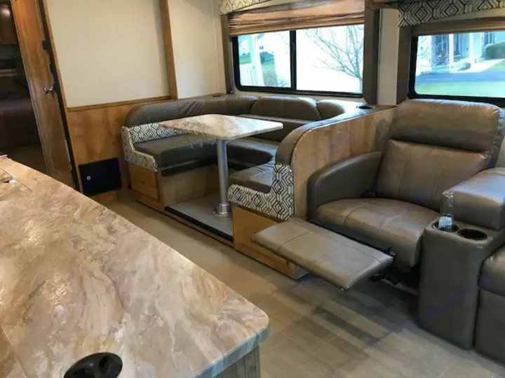 RV rental interior: A U-shaped dinette, table, and two reclining chairs with extended footrests & cup holders, plus a counter.. Coachmen Leprechaun 2018
