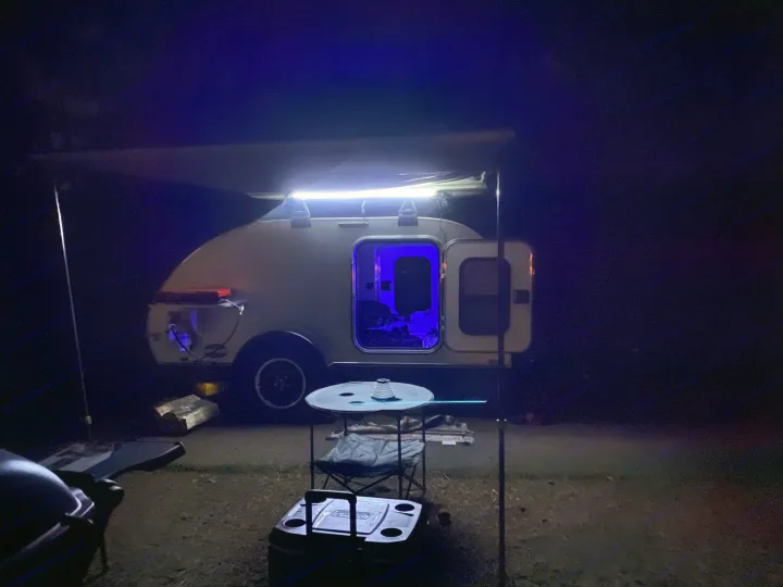 Teardrop RV rental glowing at night. White awning lights illuminate the campsite, while blue light spills from the open camper door.. Aero Teardrops Teardrop Camper 2022