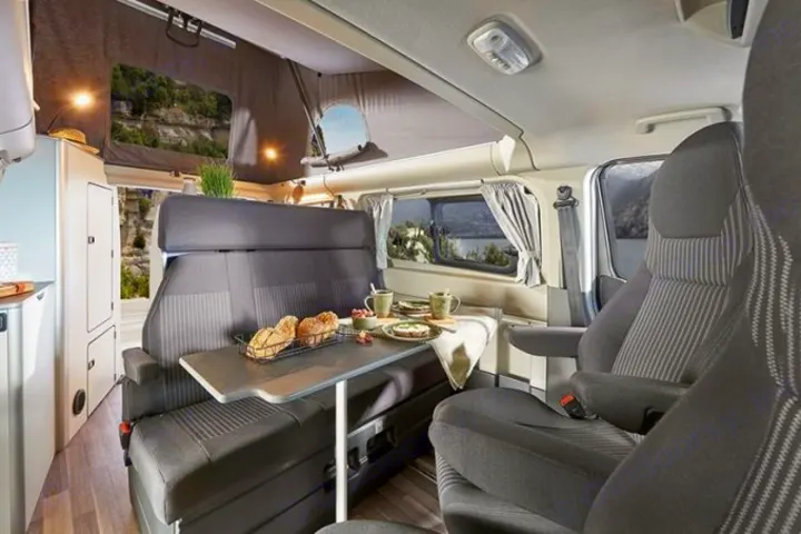 Cozy RV rental camper van interior. Dining table with food, seats, and pop-top windows reveal scenic lake views.. Ford Nugget 2021