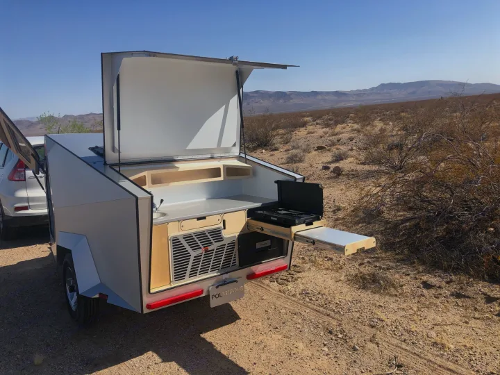 Rear view of a compact teardrop RV rental, open to reveal a full outdoor kitchen, sink, and stove in a desert setting.. Polydrop K-20 2020