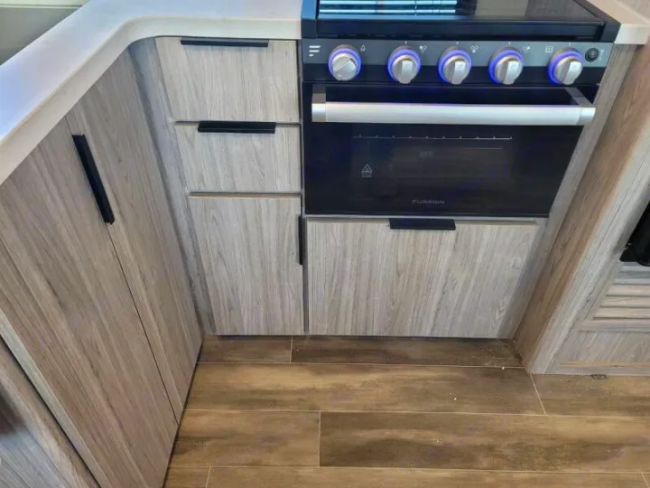 Modern RV rental kitchen: light wood-grain cabinets, white counter, black oven with blue-lit controls, and wood-look floor.. Keystone Passport 2021
