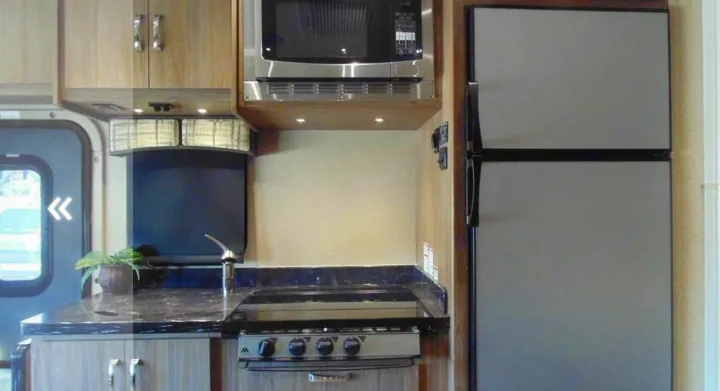 Compact RV rental kitchen featuring a microwave, refrigerator, stove, sink, and wood cabinets for your travels.. Dynamax Corp Isata 2016