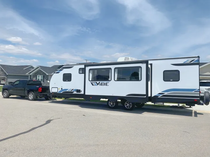 A black pickup truck tows a sleek white travel trailer RV on an asphalt road, ready for an exciting rv rental trip.. Forest River Vibe 2021