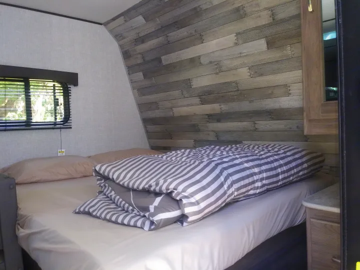 Inside an RV rental: a comfy bedroom with a grey wood accent wall, window blinds, and striped bedding. Ready for adventure.. Crossroads RV Zinger Lite 2021