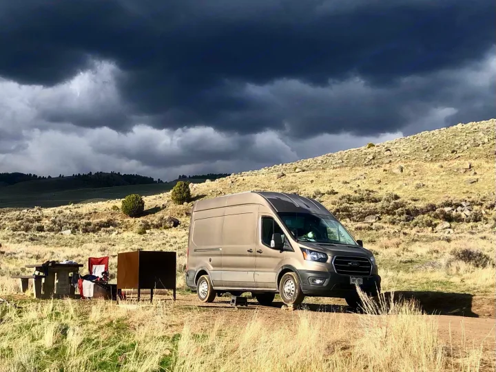RV rental: A tan camper van parked on a dirt road with a picnic setup, under foreboding dark storm clouds over dry hills.. Ford Transit 2020