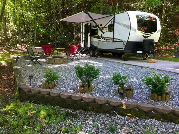 A serene RV rental campsite featuring a parked camper, gravel patio with fire pit, and seating, nestled in a wooded area.. Forest River Rockwood Mini Lite 2021