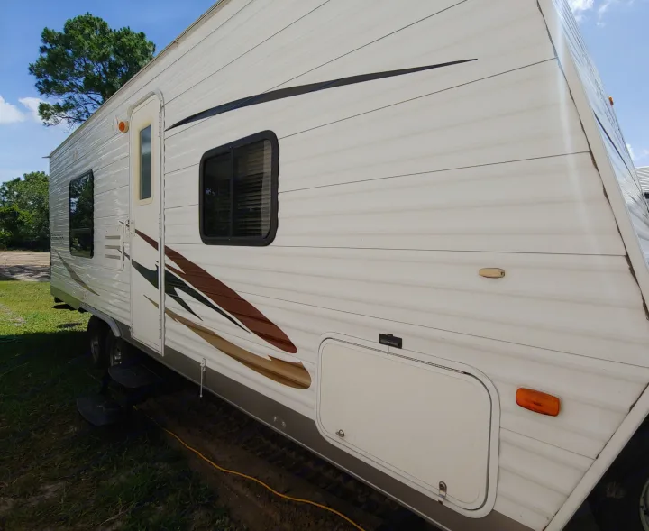 White travel trailer for an RV rental, parked outdoors on grass. Visible door, windows, and decorative stripes on its side.. Coachmen Catalina 2011